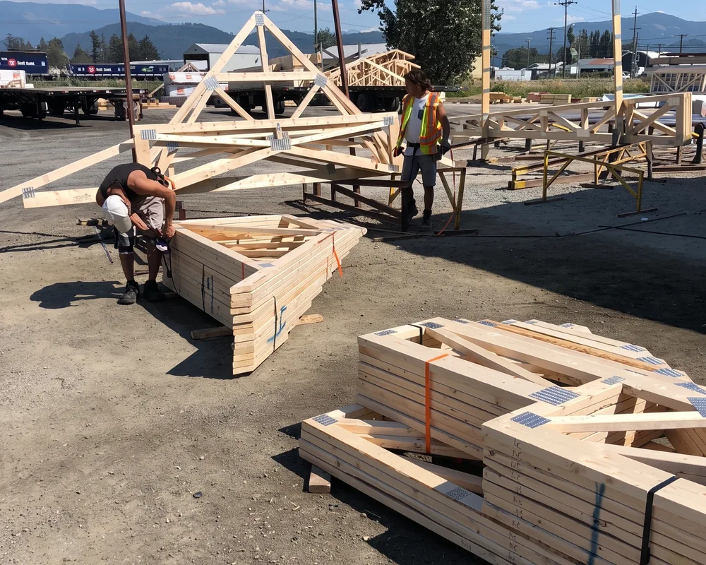 Workers assemble wooden roof trusses at an outdoor construction site with mountains visible in the background.