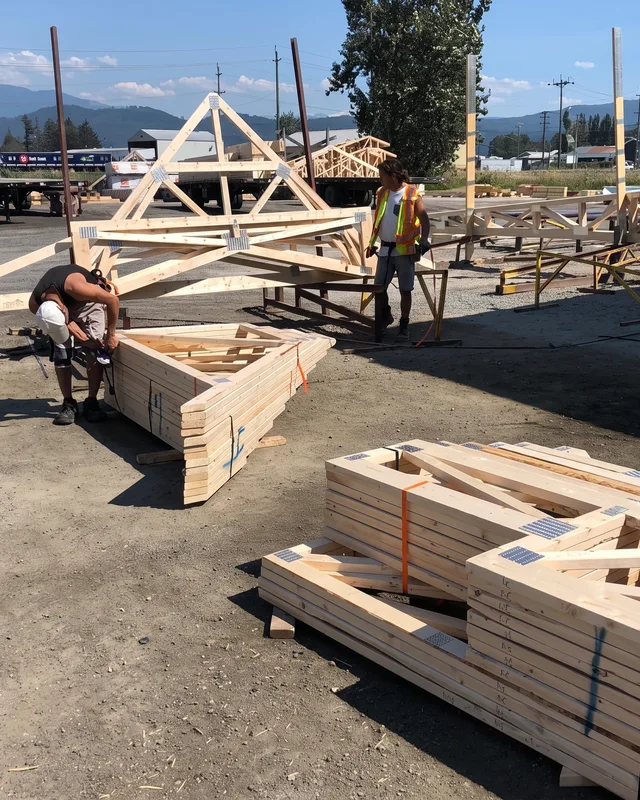 Workers assemble wooden roof trusses at an outdoor construction site with mountains visible in the background.