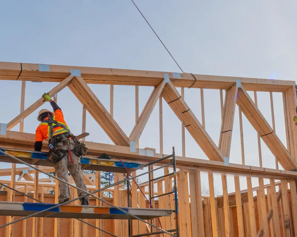 Two construction workers install wooden roof trusses on a residential home frame under blue sky.
