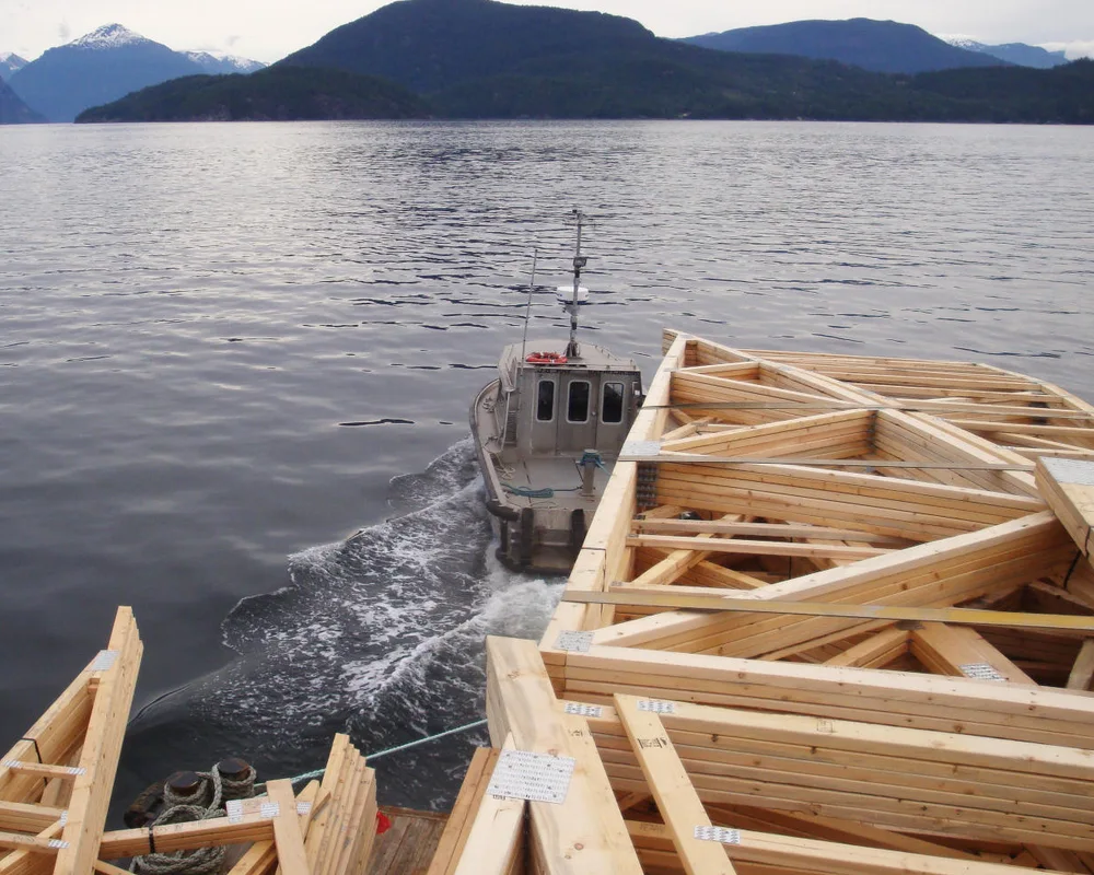 Wooden boat frames dry on a dock beside a small motorboat on calm water with mountains in the background.