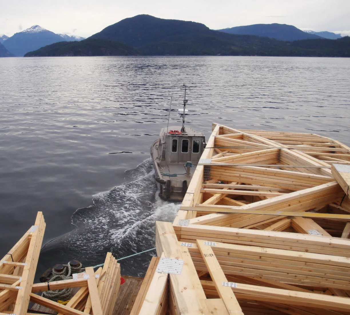 Wooden boat frames dry on a dock beside a small motorboat on calm water with mountains in the background.